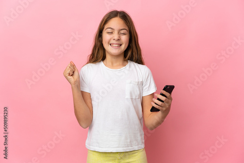 Child over isolated pink background using mobile phone and doing victory gesture