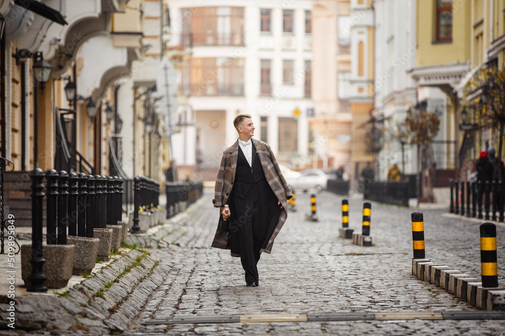 Fototapeta premium Young elegant man actor dressed in long coat and classic tailcoat on background of City. Fashion style guy with wine bottle. Street style male portrait looking to camera. Modern urban walk in town
