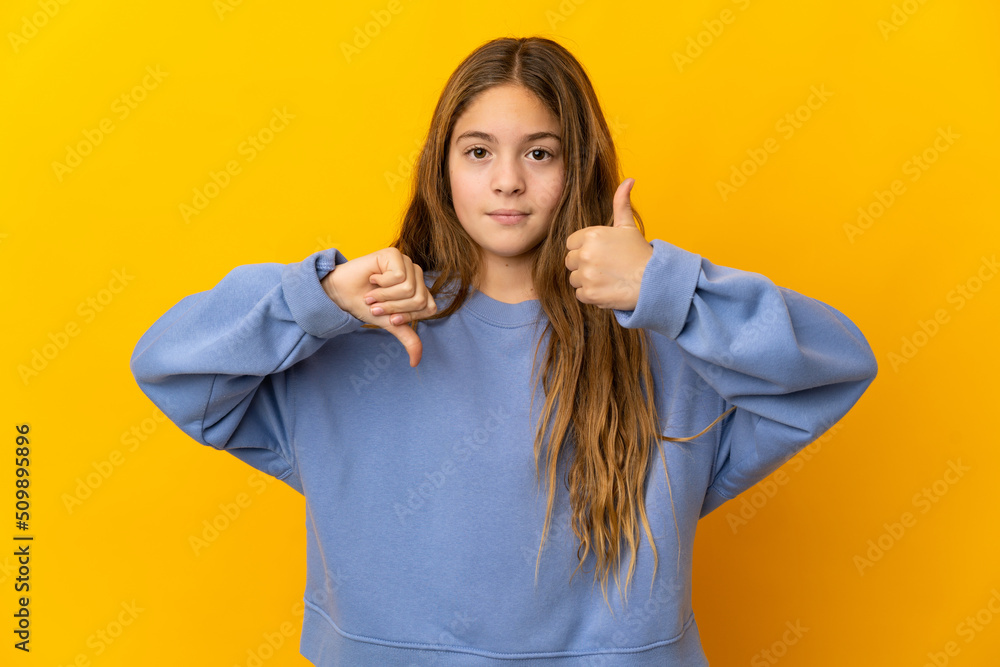 Child over isolated yellow background making good-bad sign. Undecided between yes or not Child over isolated yellow background making good-bad sign. Undecided between yes or not