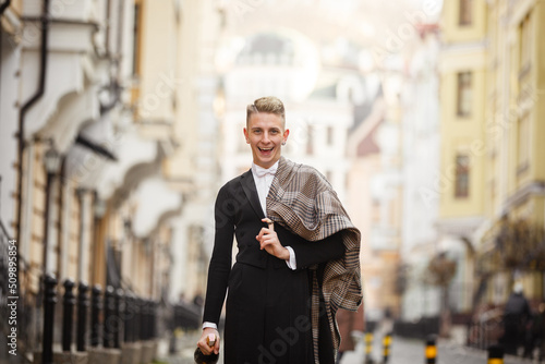 Young elegant man actor dressed in long coat and classic tailcoat on background of City. Fashion style guy with wine bottle. Street style male portrait looking to camera. Modern urban walk in town photo
