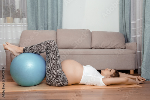 Beautiful pregnant woman at fitness gym with on aerobics ball. Outfit exercising with a pilates ball. Pregnant woman on yoga mat holding legs on transparent fitness ball in home.