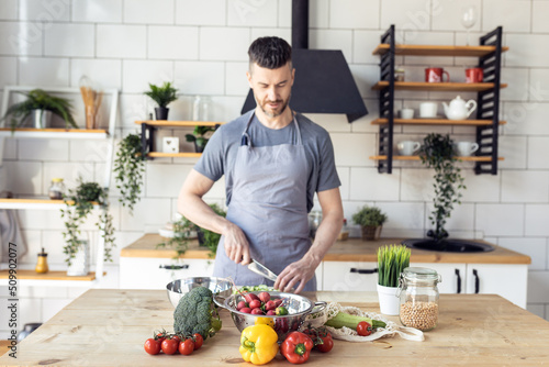 Handsome father, strong young man cooking healthy vegetable salad with fresh organic ingredients, tasty food in the kitchen at home . Men doing chores. Ripe pepper, tomato, cucumber