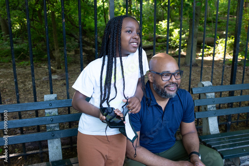 An African-American man and his young daughter sitting on a bench in the park (ID: 509903610)