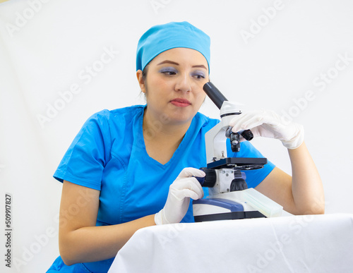 beautiful female lab technician wearing uniform and blue surgical cap, latex gloves, analyzing samples with the microscope, on white background © Liliana Kinomoto