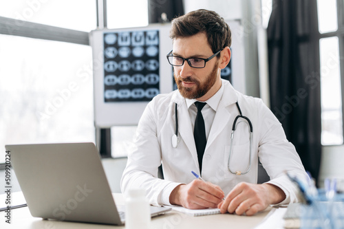 Caucasian smart doctor attentively listens to patient's complaints via video link, while sitting at workplace, writes out a prescription to the patient, prescribes treatment, makes recommendations