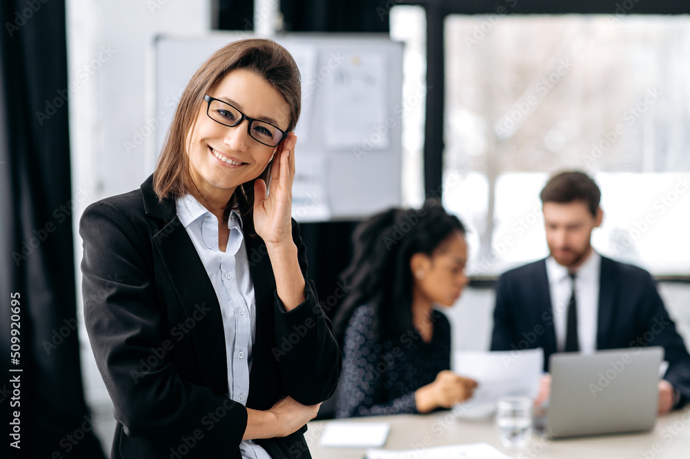Positive confident elegant gorgeous caucasian business woman with glasses, in a formal suit, creative director, business leader, standing in a modern office, looking at the camera, smiling friendly Positive confident elegant gorgeous caucasian business woman with glasses, in a formal suit, creative director, business leader, standing in a modern office, looking at the camera, smiling friendly