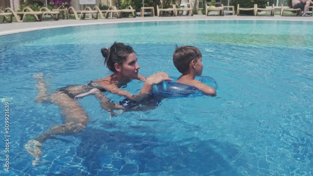 Mother teaches her son swiming in a public pool. The child is swinging in lifebuoy on an artificial wave in the tropical spa. Mother teaches her son swiming in a public pool. The child is swinging in lifebuoy on an artificial wave in the tropical spa.