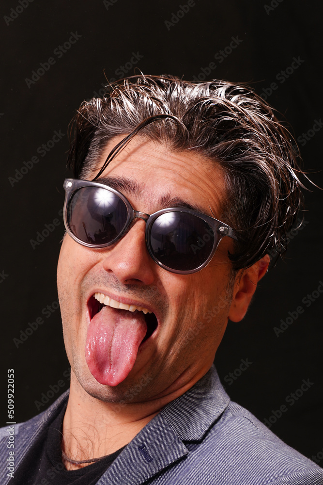 Smiling young man sticking out his tongue looking at the camera with black sunglasses, wearing a black T-shirt and a blue jacket, on black background. Smiling young man sticking out his tongue looking at the camera with black sunglasses, wearing a black T-shirt and a blue jacket, on black background.