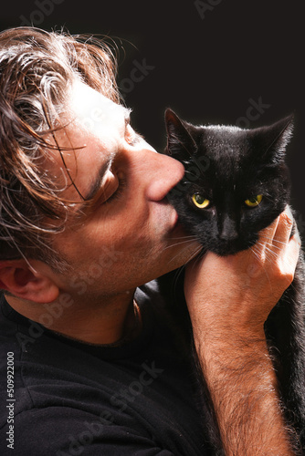  young man kissing a black cat with yellow eyes, wearing black t-shirt on black background.