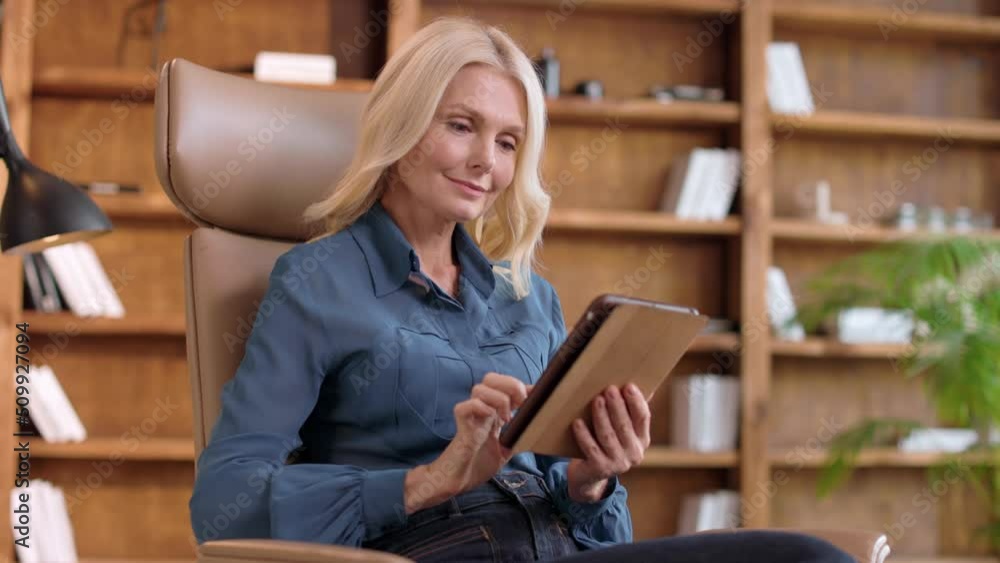 Positive female psychologist using digital tablet in brown chair with with wooden shelves on blur background. Beautiful senior woman with blond hair planning schedule for next week. Positive female psychologist using digital tablet in brown chair with with wooden shelves on blur background. Beautiful senior woman with blond hair planning schedule for next week.