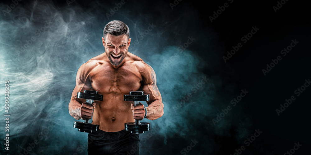 Adult muscular man screaming while working out hard with dumbbells on dark studio background with smoke