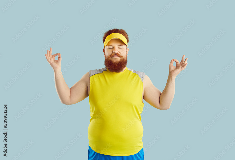 Naklejka premium Funny yoga. Funny fat man with serious facial expression meditating isolated on light blue background. Young redhead man in sports uniform and with big belly doing yoga with his eyes closed.