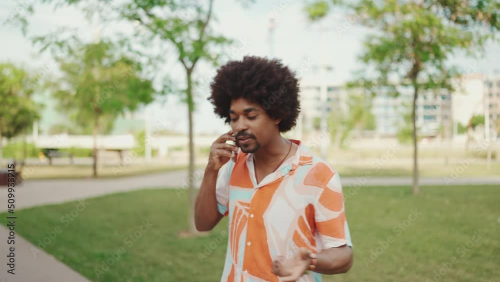 Close-up of young African American man wearing shirt walking and talking on smartphone on path in the park. Man emotionally talking on mobile phone. Lifestyle concept. Slow motion Close-up of young African American man wearing shirt walking and talking on smartphone on path in the park. Man emotionally talking on mobile phone. Lifestyle concept. Slow motion