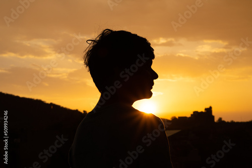 Panoramic silhouette shot of an Indian kid looking at the orange sky during the sunset. Hopeful kid stares at the sun. Kid lost in deep thoughts after looking at the sunset. 