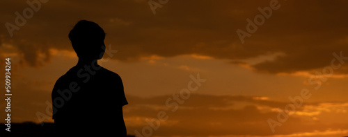 Panoramic silhouette shot of an Indian kid looking at the orange sky during the sunset. Hopeful kid stares at the sun. Kid lost in deep thoughts after looking at the sunset. 