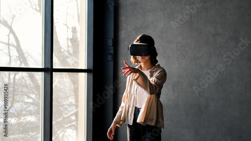 A teenage girl in virtual reality glasses stands in a gray room near a large window.
