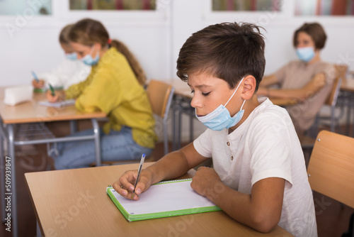 Obraz na plátně Portrait of schoolboy in protective face mask sitting at desk in classroom durin