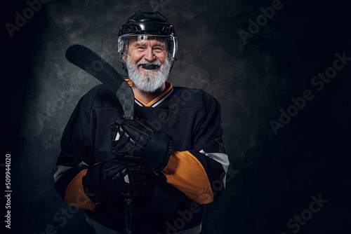 Portrait of happy elderly sportsman dressed in black protective sportswear holding hockey stick.
