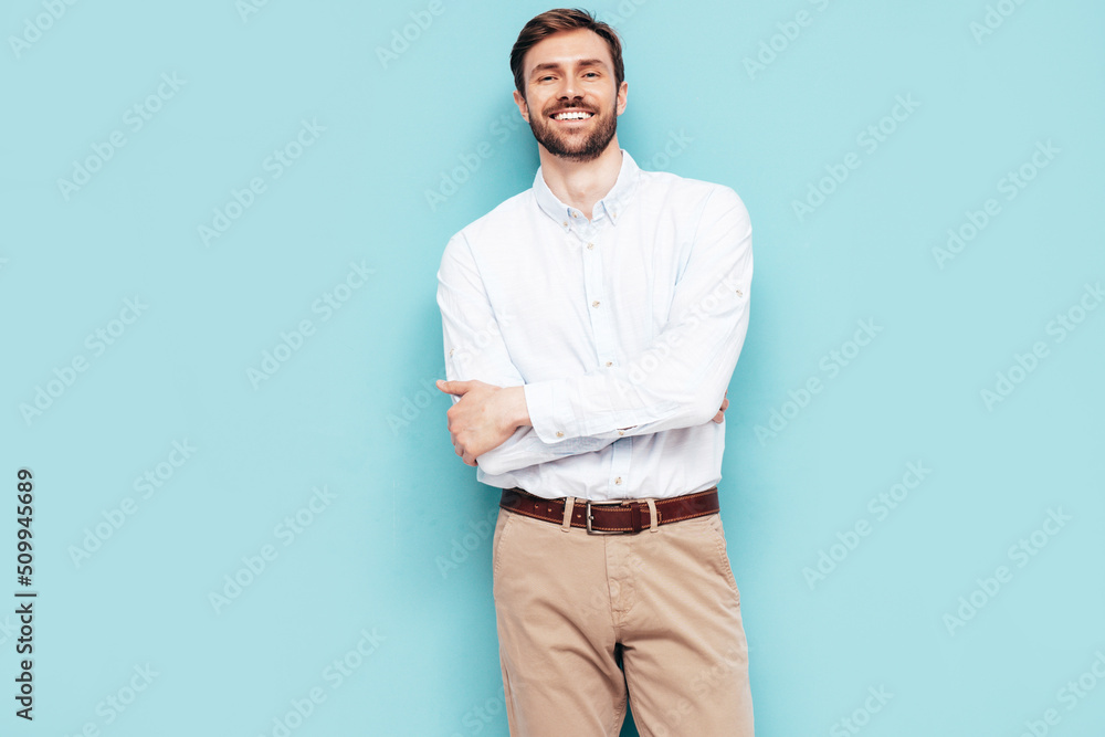 Portrait of handsome smiling model. Sexy stylish man dressed in shirt and trousers. Fashion hipster male posing near blue wall in studio. Crossed arms Portrait of handsome smiling model. Sexy stylish man dressed in shirt and trousers. Fashion hipster male posing near blue wall in studio. Crossed arms