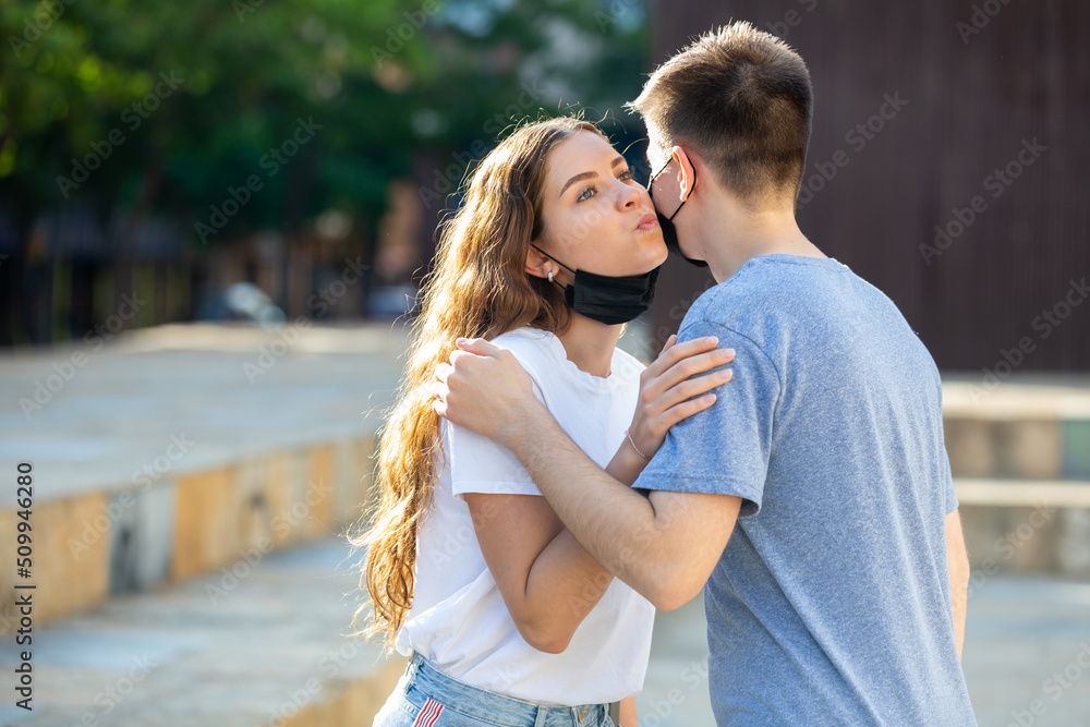 Boy and girl in protective masks kiss each other on cheek on the street. High quality photo