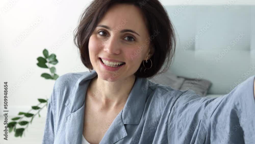 Smiling european female sitting  on sofa hold smartphone talk by video call wave hand to camera. Positive young mixed race woman contact call friend by video link chat using device webcam