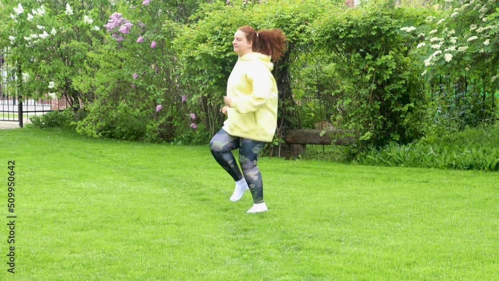 Red-haired smiling curly fat woman with excess weight in yellow hoodie do sports exercises, jumping, high knee taps, jogging in place. Green backyard or park on background. Weight loss in summer Red-haired smiling curly fat woman with excess weight in yellow hoodie do sports exercises, jumping, high knee taps, jogging in place. Green backyard or park on background. Weight loss in summer