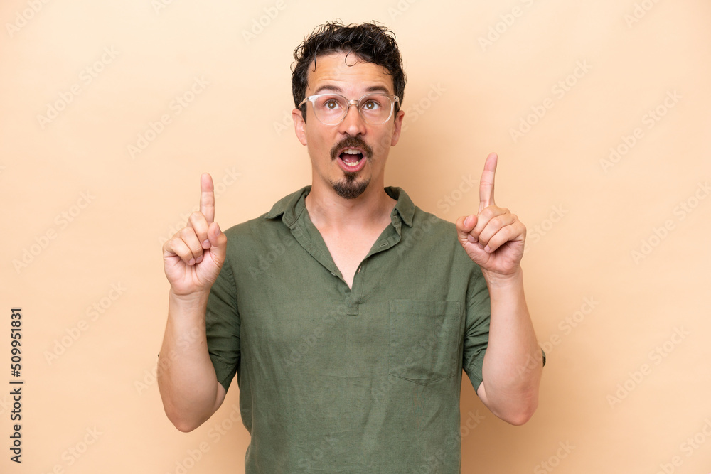 Young caucasian man isolated on beige background surprised and pointing up Young caucasian man isolated on beige background surprised and pointing up