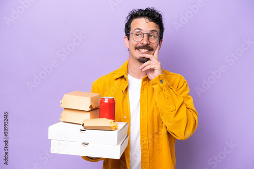 Young caucasian man holding fast food isolated on purple background thinking an idea while looking up