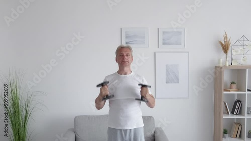 Home training. Morning workout. Weight lifting. Strong athletic middle-aged man doing physical exercise for arm muscles with dumbbells in light modern interior. photo