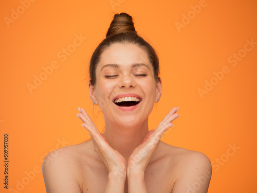 Face portrait of smiling woman with open mouth and closed eyes, bun hair stule and bare shoulders. Hands in frame near face. photo