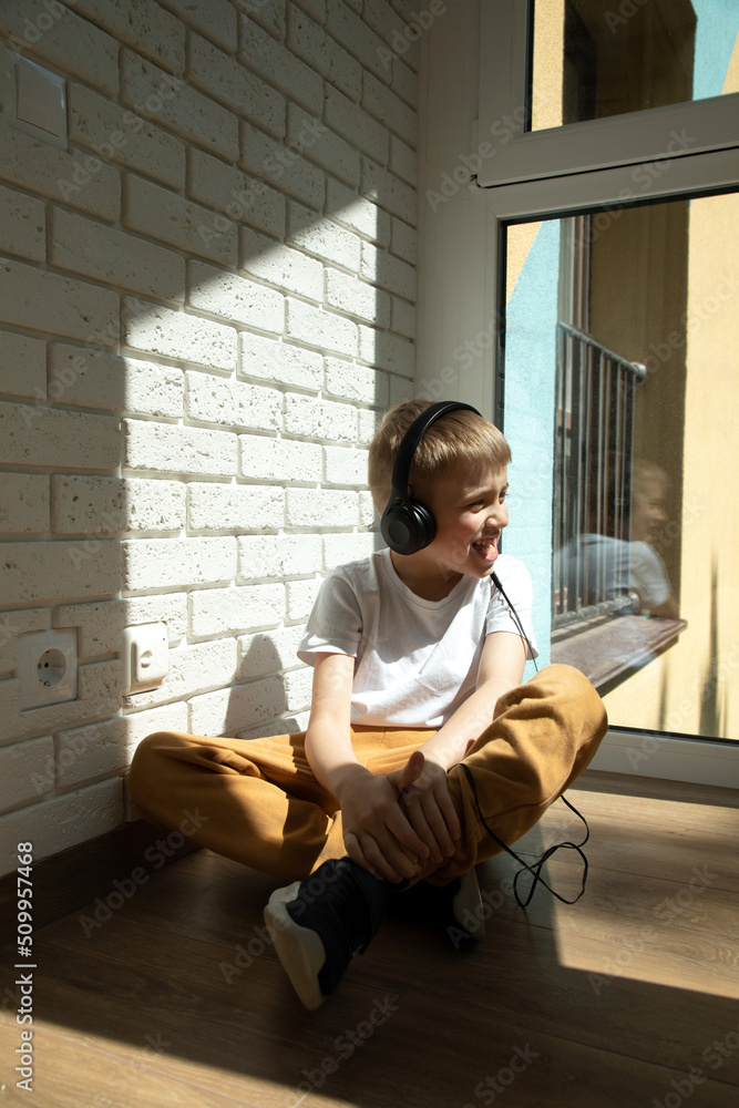 A boy of 6-7 years old in a white T-shirt listens to music in black headphones, sings and looks out the window