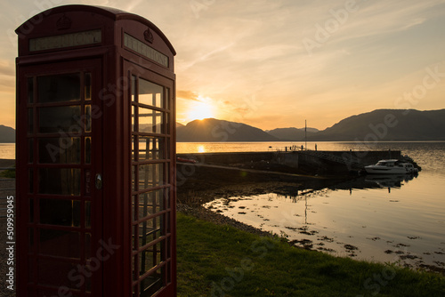View of Loch Leven under a  dramatic red sky at sunset in Glencoe, Scottish Highlands, UK, with a red Phone booth on the foreground and a yacht next to a pier on the background