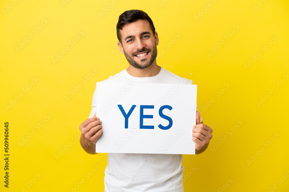 Young handsome caucasian man isolated on yellow background holding a placard with text YES with happy expression Young handsome caucasian man isolated on yellow background holding a placard with text YES with happy expression