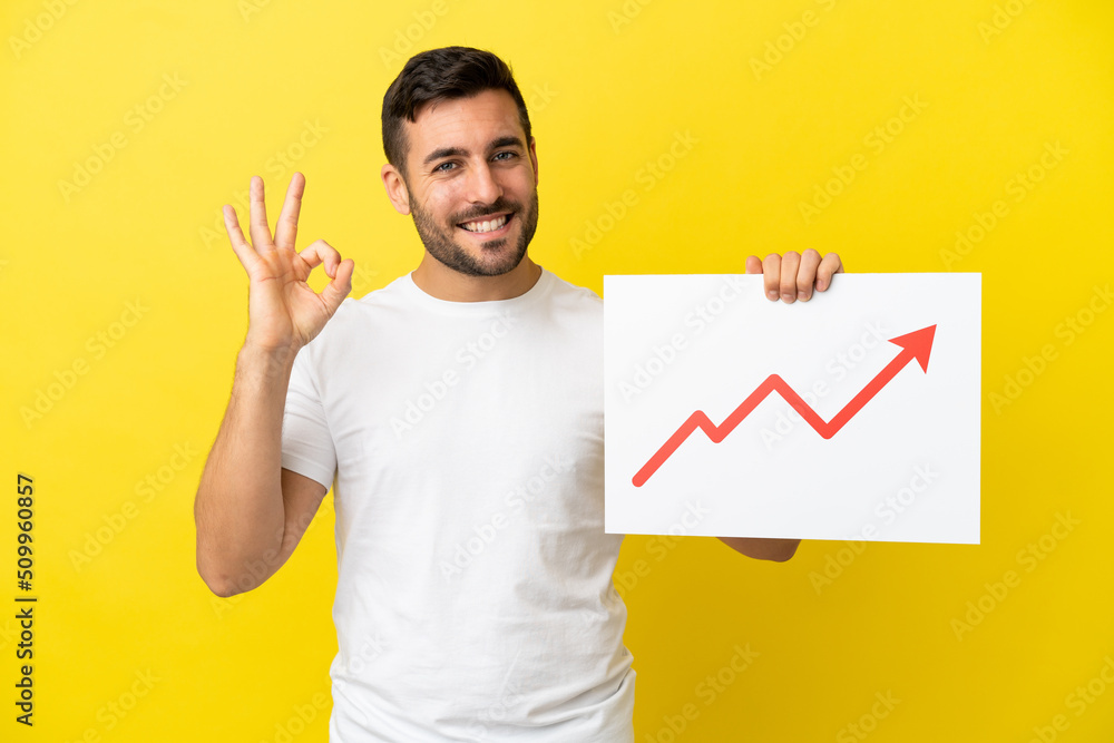 Young handsome caucasian man isolated on yellow background holding a sign with a growing statistics arrow symbol with ok sign Young handsome caucasian man isolated on yellow background holding a sign with a growing statistics arrow symbol with ok sign