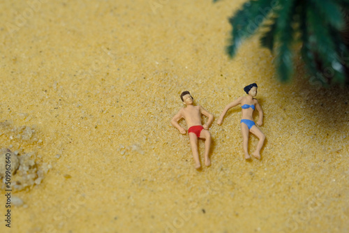 Miniature people toy figure photography. Top eagle view of men and girl couple relaxing, lying at beach sand when daylight at seaside