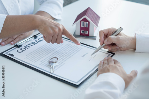 A real estate agent with a house model is talking to clients about buying home insurance and having customers sign contracts under the formal contract agreement. Home rental and insurance concept