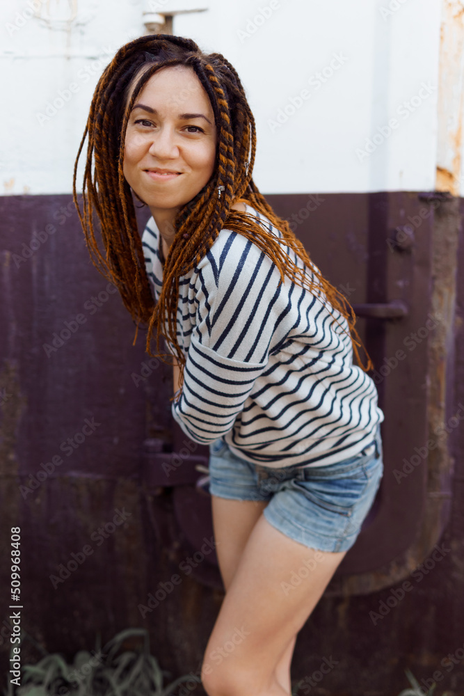 Naklejka premium a girl with a dreadlocked hairstyle poses in the summer outdoors, dressed in a T-shirt and denim shorts