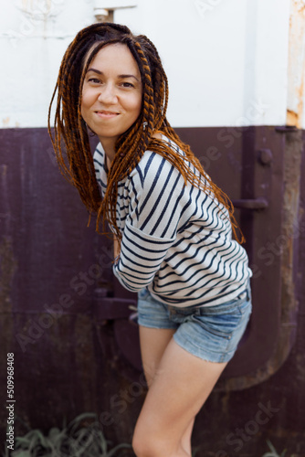 a girl with a dreadlocked hairstyle poses in the summer outdoors, dressed in a T-shirt and denim shorts