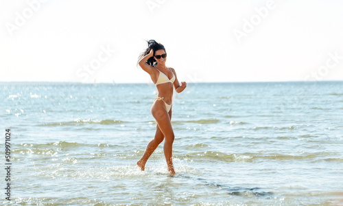 people, summer and swimwear concept - young woman in bikini swimsuit running in shallow water on beach