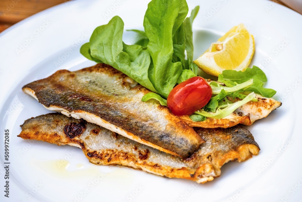 Rousted trout fillet with salad leaf, tomato and lemon on white plate.