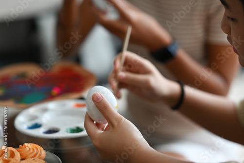 Side view close up with Asian female kid holding egg for painting Easter holiday symbol with her older sister over blurred color palette background.