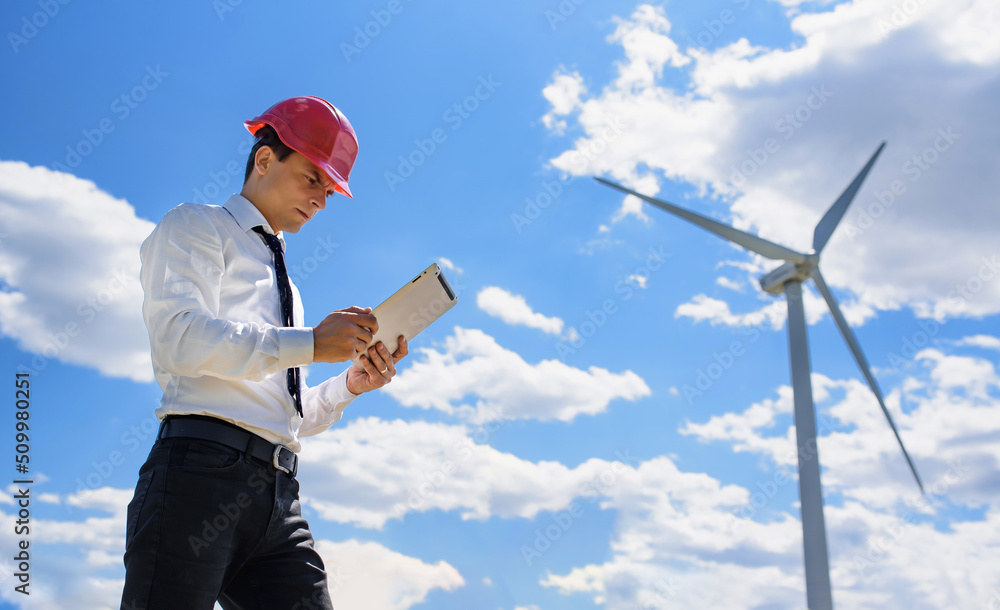 Young man engineer in white shirt and red safety helmet holding a tablet and verifying the wind turbine windmill. Young man engineer in white shirt and red safety helmet holding a tablet and verifying the wind turbine windmill.