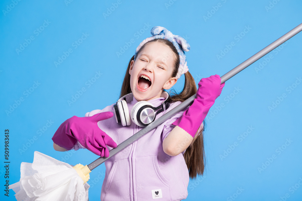 Beautiful young girl with headphones and mop sings on blue background. Pretending to play guitar with rubber pink gloves, she has wireless headphones. Beautiful young girl with headphones and mop sings on blue background. Pretending to play guitar with rubber pink gloves, she has wireless headphones.