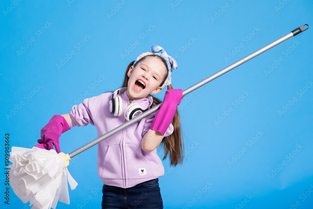 Beautiful young girl with headphones and mop sings on blue background. Pretending to play guitar with rubber pink gloves, she has wireless headphones. Beautiful young girl with headphones and mop sings on blue background. Pretending to play guitar with rubber pink gloves, she has wireless headphones.