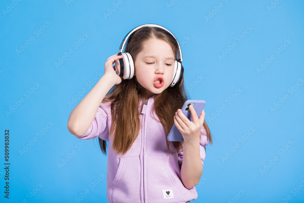 Beautiful little girl with long brunette hair with phone listening to music through wireless isolated headphones on blue background. Joyful mood of young child enjoying a song.