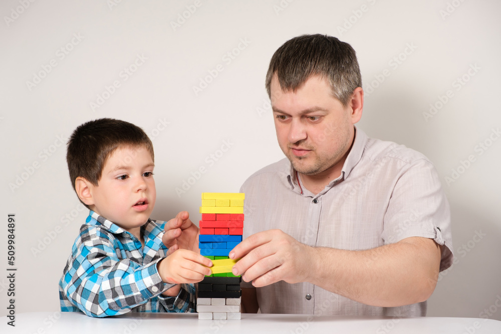 Father and son together build a tower of colorful wooden blocks sitting at a table Father and son together build a tower of colorful wooden blocks sitting at a table