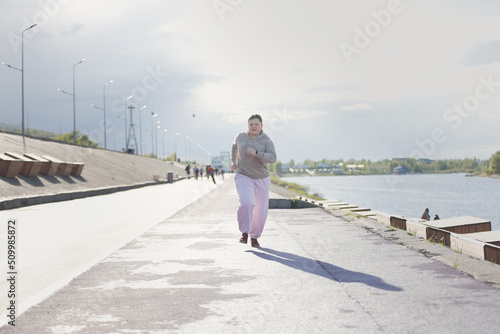 overweight European teenage girl in tracksuit jogging along concrete embankment, outskirts of city. Sports and adolescents, © natalialeb