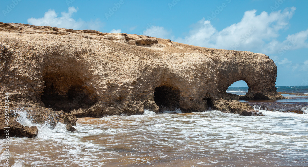 The seashore with a stone hill and washed round holes openings caves against the background of water and blue sky on a clear sunny day. Nature ocean landscape ecology.