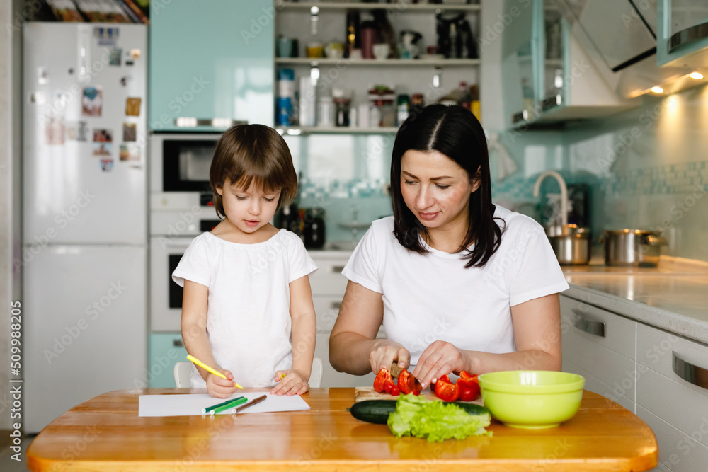 Mother and little daughter sitting in the kitchen, making vegetable salad, drawing. Happy family concept. Daily home life Mother and little daughter sitting in the kitchen, making vegetable salad, drawing. Happy family concept. Daily home life