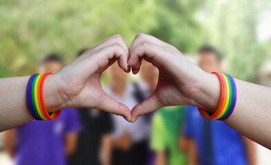 Fingers Heart shape with rainbow wristband and blurred asian teenager background, lgtbq+ celebrations concept.